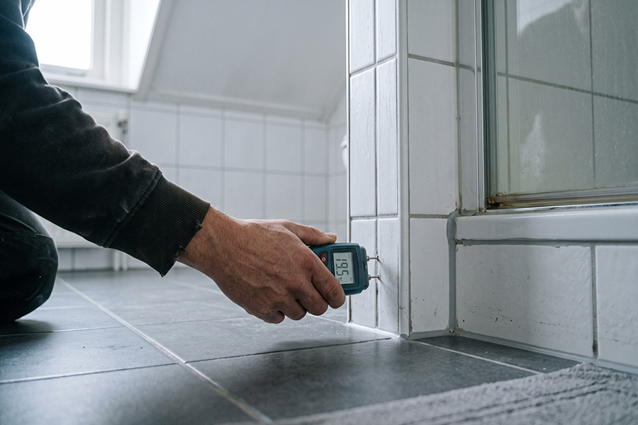 Hand met een vochtmeter tegen de voeg onder aan een douchewand
